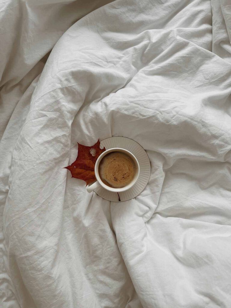 A cup of coffee on a saucer with a dried autumn leaf beside it, placed on a bed with crumpled white sheets, creating a cozy, serene fall morning vibe.