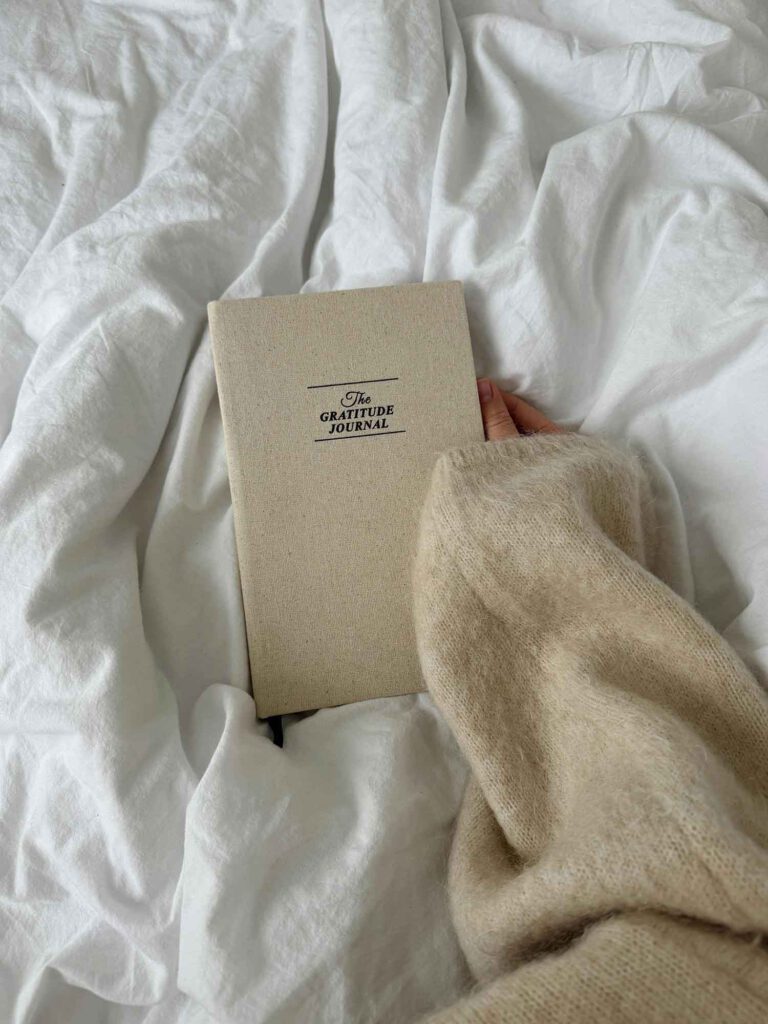 A cozy scene featuring a gratitude journal resting on white bedding, held by a hand in a soft beige sweater.